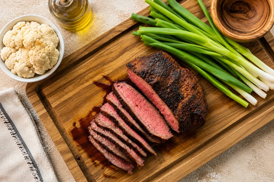 A Blackened Santa Maria Tri-Tip steak served on a wooden board with fresh scallions, bowl of cauliflower, and oil on the sides.